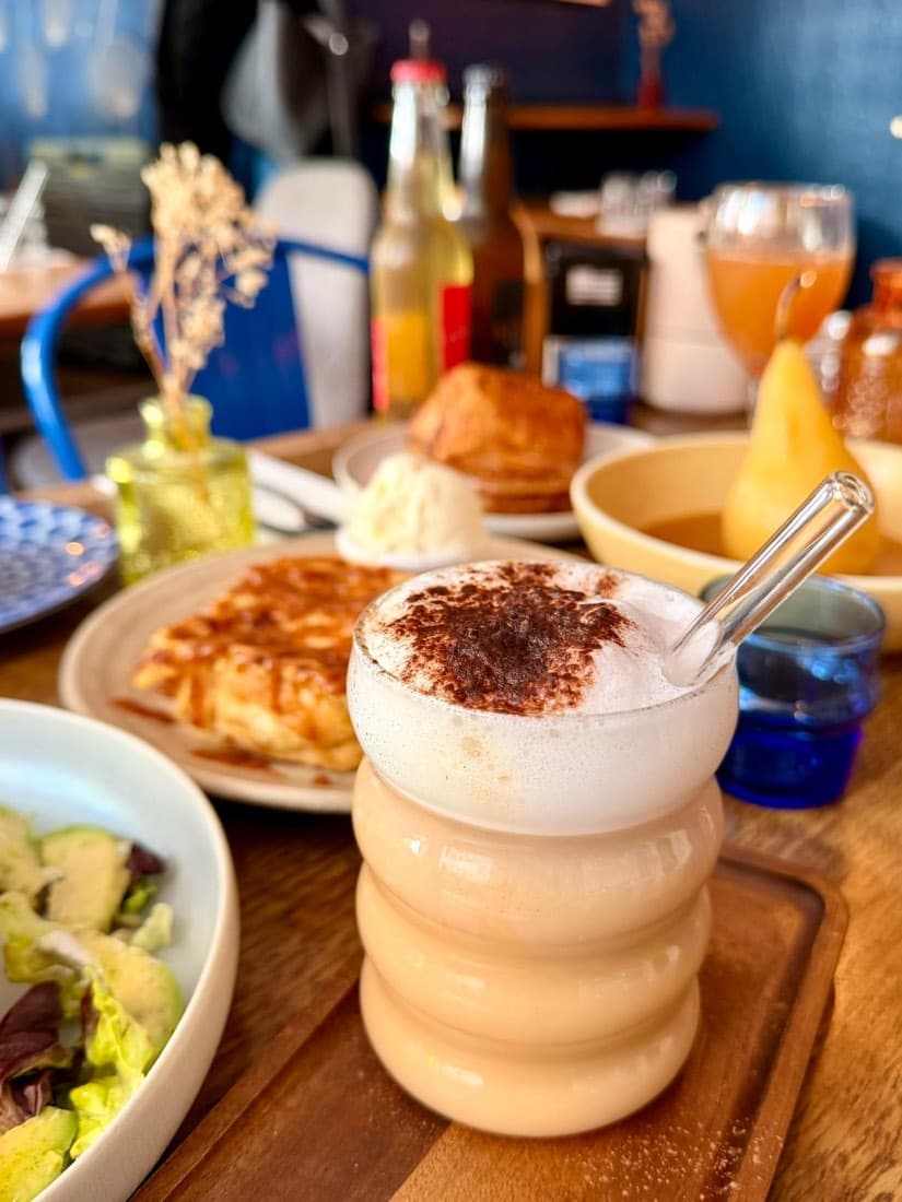 Un latte mousseux saupoudré de cacao, servi dans un verre ondulé au brunch de Ty Louis