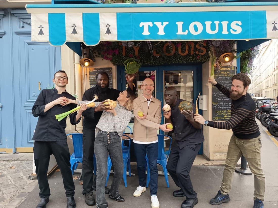 Toute l’équipe de Ty Louis pose en souriant avec des légumes bretons devant la devanture bleue du restaurant, rue du Faubourg Saint-Antoine à Paris 12