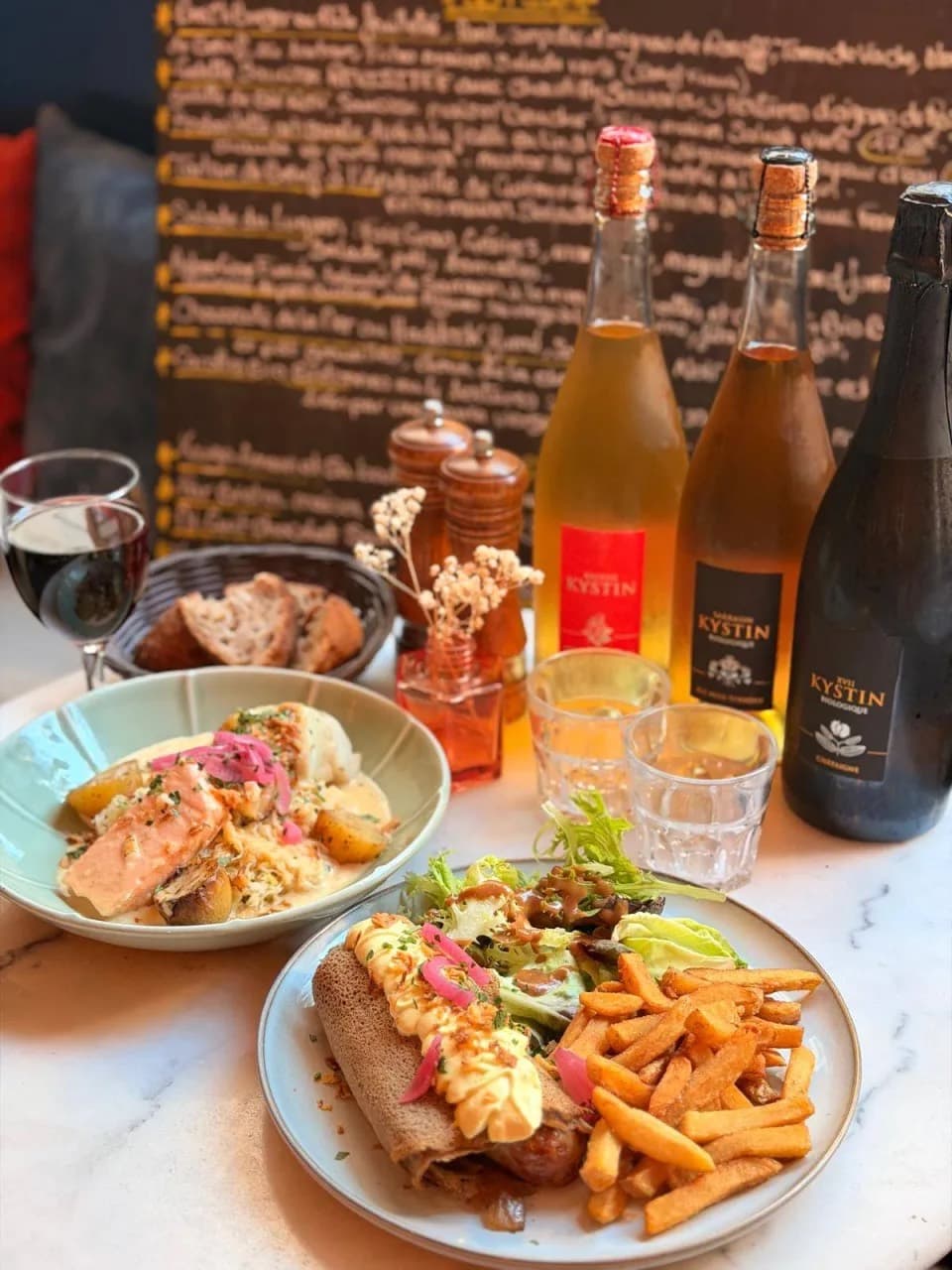 Deux plats de la Traversée bretonne, la choucroute de la mer et la galette-saucisse revisitée avec ses frites, devant trois bouteilles de cidre Kystin chez Ty Louis
